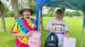 Two men stand under a gazebo tent holding backpacks and a binder. Ronald Piaseczny, on the left, is wearing a grey bucket hat and rainbow tie-dye shirt. Mark Smith, on the right, is wearing a baseball cap, glasses, and a shirt with the Niagara Pride logo on it.