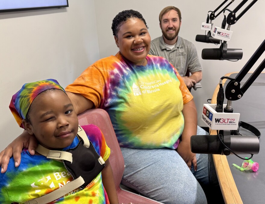 Three people sit in a radio studio. A smiling woman and a man sit behind a child wearing a tie-dye headscarf and shirt, who is winking. All are near microphones labeled WGLT.