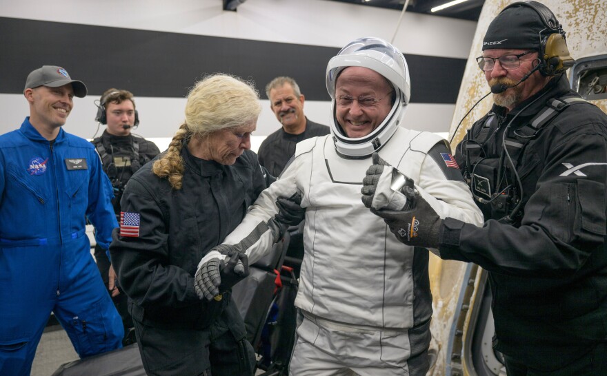 Man in white spacesuit and helmet smiles as he's helped by two people in dark, warm weather uniforms. Other people stand in the background.