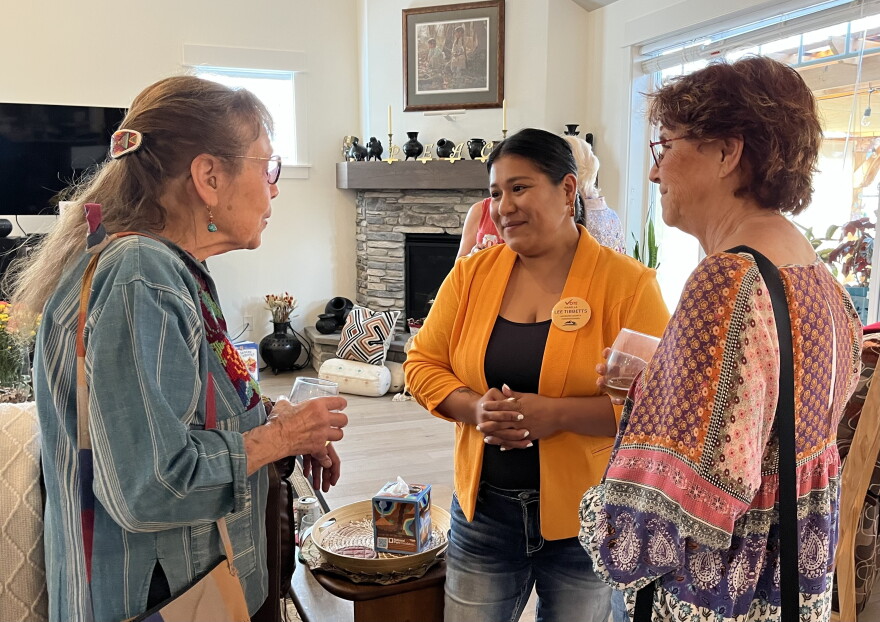 Three women are standing in a circle and talking. The woman in the center is wearing an orange blazer and has her hands clasped in front of her. 