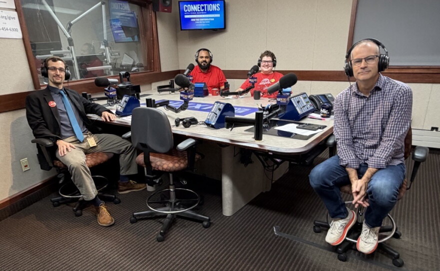 Four smiling people wearing headphones sit at a table in a radio talk studio: a man front left has short brown hair and a brown mustache and beard and is wearing glasses, a dark blue button-down shirt and lighter blue tie under a black suit coat, khakis, dark socks, and brown shoes; a man back left has short black hair and a black mustache and beard and is wearing a red polo shirt; a woman back center has short brown hair and is wearing glasses and a red graphic t-shirt; a man at right has very short dark hair and is wearing glasses, a purple and white checked button-down shirt, jeans, and white and red sneakers