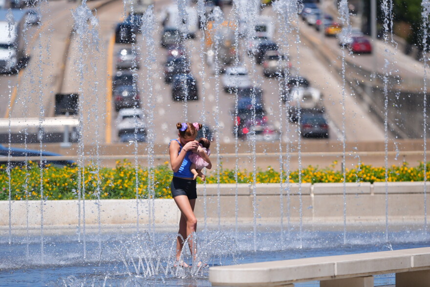 A young girl with brown hair in shorts and a tanktop holding a doll walks through a fountain that overlooks a highway.
