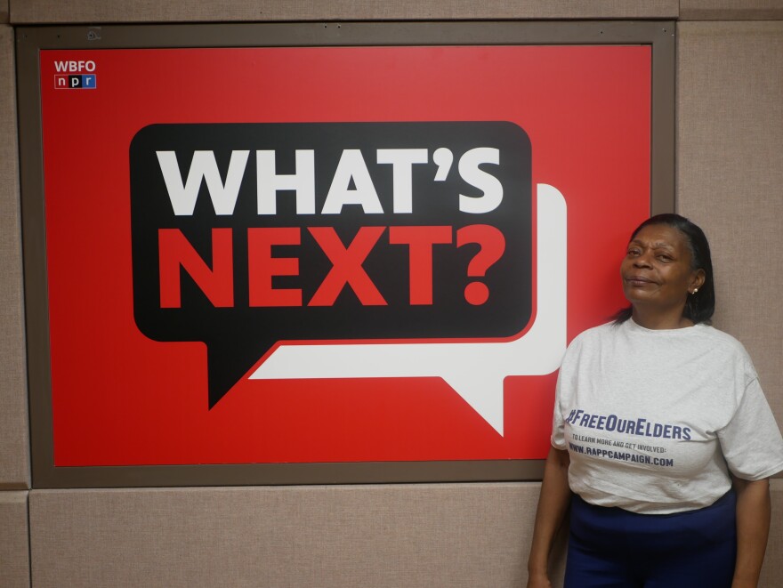 A woman in a white shirt stands in front of a red black, and white What's Next? sign