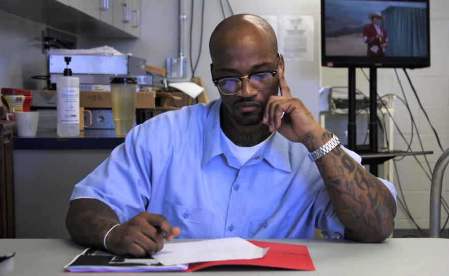 A Black man with glasses in blue prison clothes looks down at a love letter on a desk, with a contemplative look. 
