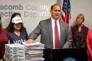 Macomb County Clerk and Republican candidate for Michigan secretary of state Anthony Forlini speaks at a podium in a county elections office. He stands before stacks of documents and boxes of copy paper, flanked by several people. American and Michigan flags stand behind him, with election-office signage on the wall.