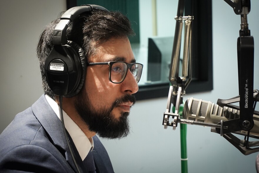 A man wearing glasses talks into a microphone in a radio studio.