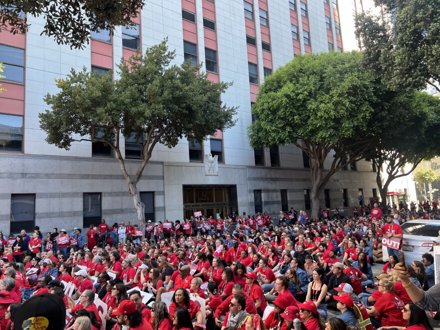 A crowd of more than a thousand nurses wearing red rally in front of a large building with grey cinderblocks and verticle windows, blocking the entire street and sidewalk. 