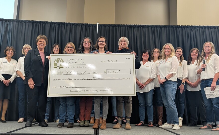 A group of women stand on a stage with black curtain in background. They are holding a large check made out to KAXE.