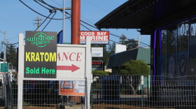 A strip mall during the day. A sign reads "Kratom Sold Here" outside of a smoke shop in a strip mall.