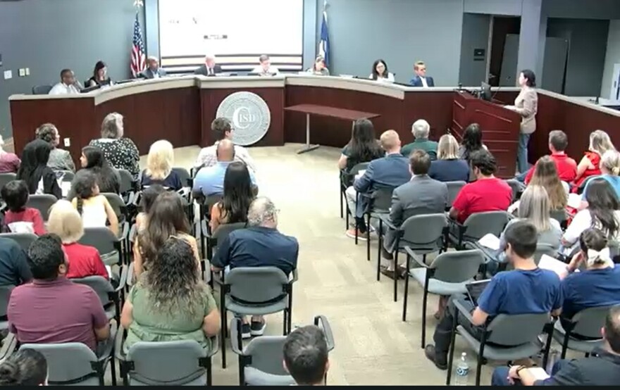 A school board meeting, from behind the seated audience, facing trustees and a speaker