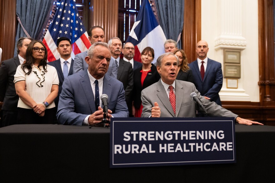 Robert F. Kennedy Jr. sits next to Gov. Greg Abbott as he speaks behind a sign that says "Strengthening Rural Healthcare."