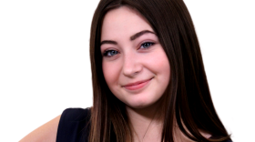 A young woman with long brown hair stands in front of a white background with her hands on her hips. She wears a black dress and smiles