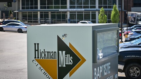 A square box lies in the foreground. On one side, it reads "Hickman Mills C-1 Schools." A large brick building with a glass front reads "Administration Center" behind it. There are several cars parked in the parking lot in the foreground.
