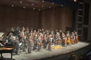 An orchestra all wearing black facing the audience on a grey stage.