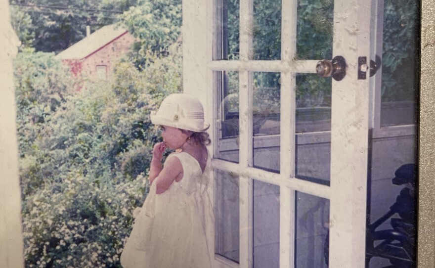 A young girl dressed in white is stepping her foot outside the door of a beautiful garden of white flowers.