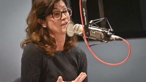 A woman sitting inside a radio studio talks at a microphone and gestures with her right hand.
