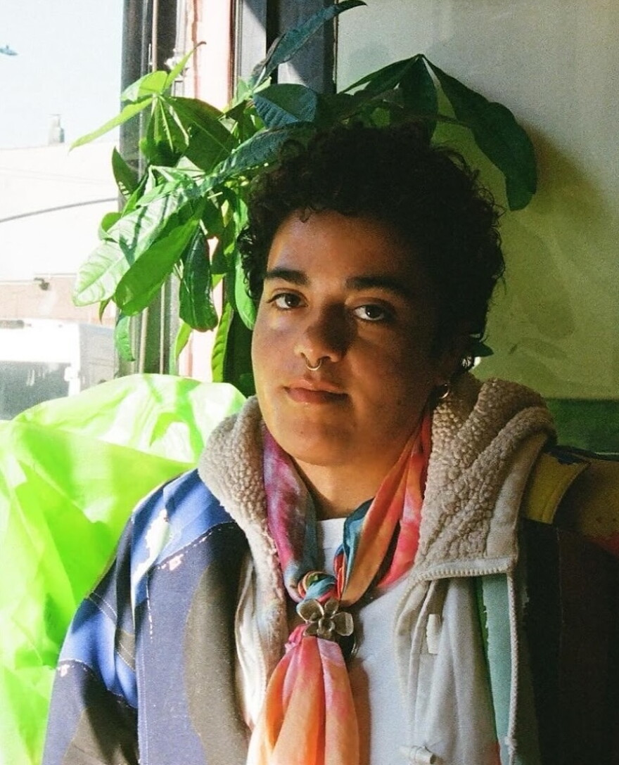 person with short curly hair wearing a jacket and looking towards the camera