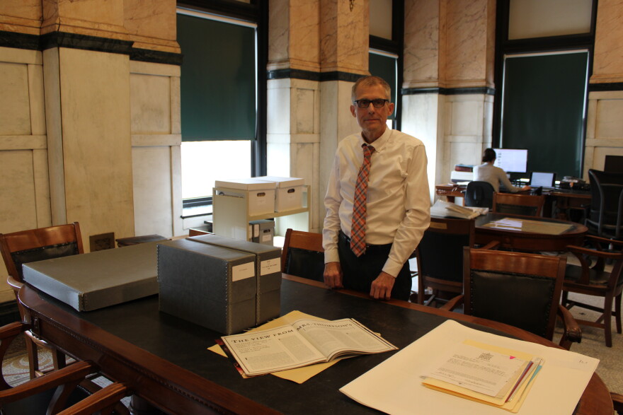 A man stands in a library surrounded by papers. In front of him is an open magazine. 