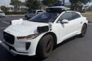 A white Jaguar car with black sensors on the front sides and on the roof sits in a parking lot in-between spots.