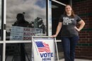 A woman in a grey shirt and jeans stands with her hand on her hip and her other on a sign that says "Vote Here." Her shirt says "Vote like your rights depend on it."