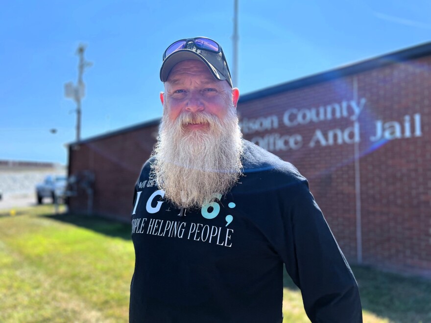 Dirk Harkins poses for a portrait. He has a long beard and wears a baseball hat and black t-shirt.