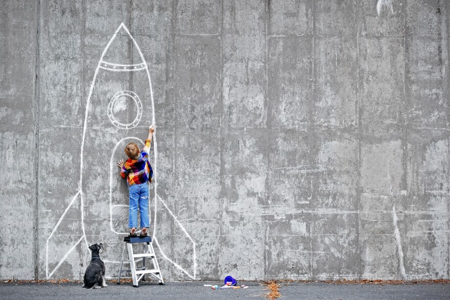 A child draws a rocket on a concrete wall with chalk as a dog looks on.