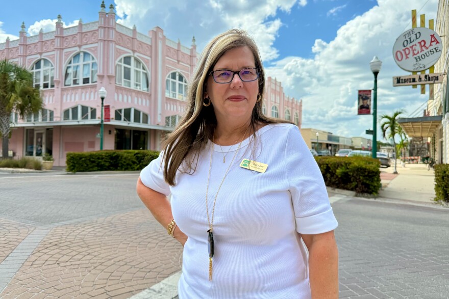 Woman with blonde hair and glasses, wearing a blue blouse and standing on a brick road with an old building in the background