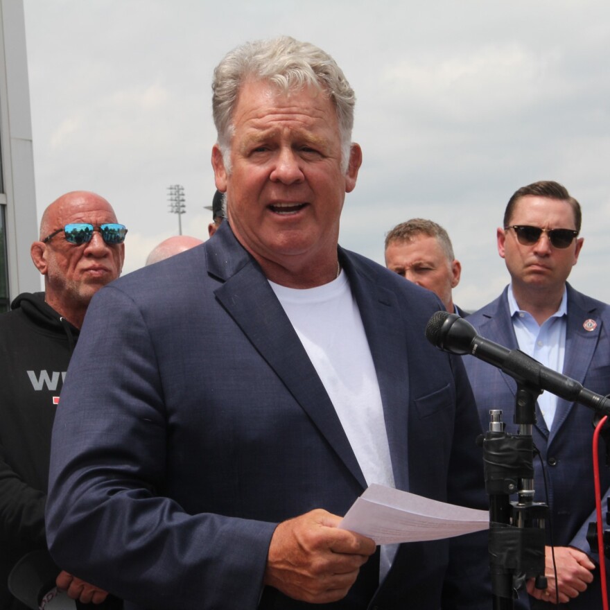 A man speaks in front of a microphone outside. A few other men stand behind him. 