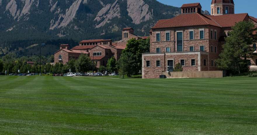 Tall brick buildings behind a large green field and the foothills of the Rocky Mountains.