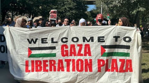 A group of people marching holding a white sheet that says Welcome To Gaza Liberation Plaza flanked by two Palestinian flags.