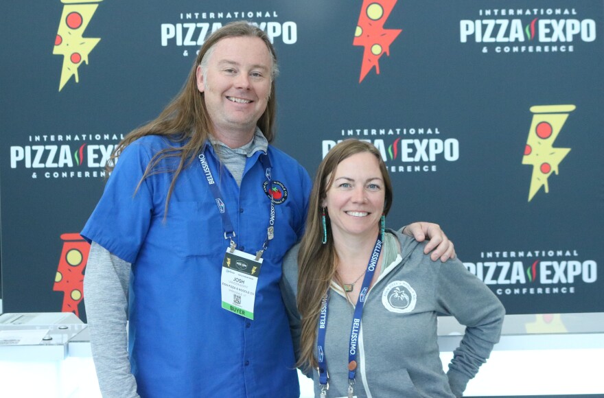 Two people in front of a large pizza expo background. 