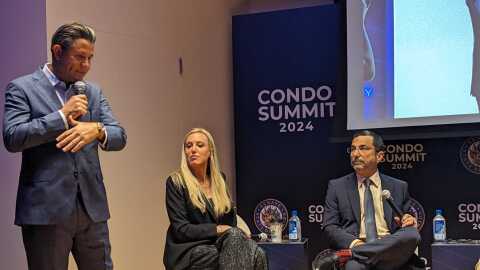 A man on the left with a microphone in a blue suit talks to a pair of panelists