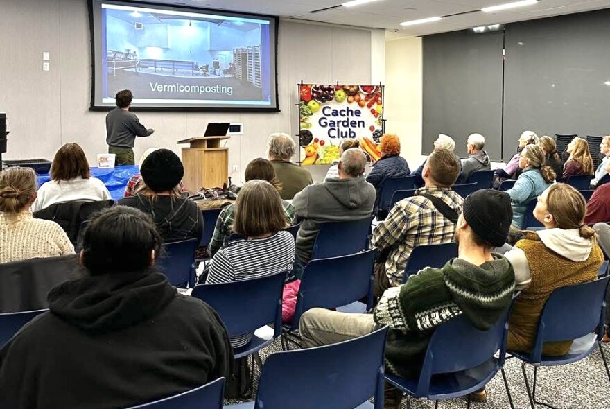 A room full of people are watching a speaker present a slideshow titled "Vermicomposting." Off to the side of the speaker, a colorful sign reads "Cache Garden Club." 