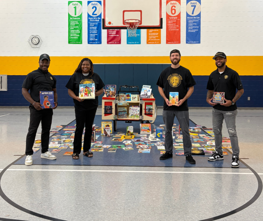 Four adults standing in a school gym holding children's books, next to a mobile book cart and dozens of books spread on the floor.
