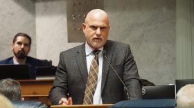 Sen. Scott Baldwin is wearing a plaid suit and a brown tie. He is speaking in the senate chambers at the statehouse. 