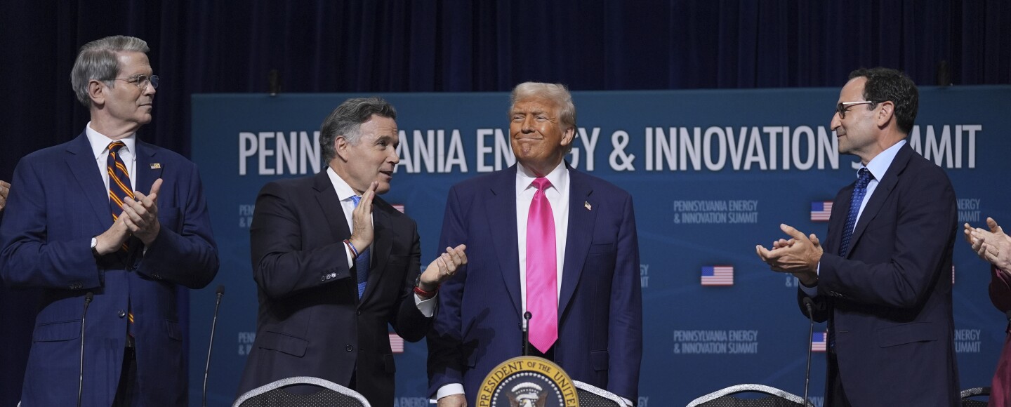 A white man in a suit smiles while standing behind a table with the presidential seal on it as three other white men look at him and applaud.