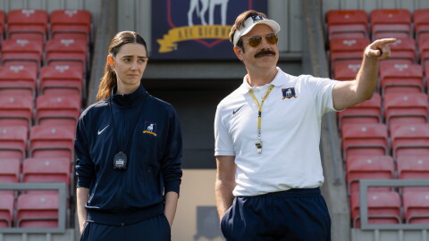 A woman and man in soccer team apparel standing in front of empty stadium seats as they look out at a soccer pitch.