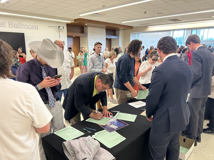 A photo of people signing green papers, registering to speak in a large hall outside an auditorium at UT Arlington.