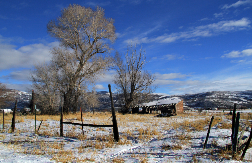 An old, worn-down shed sits in a field with yellow grass and snow. There are old wood fences in the foreground and a mountain range in the background.