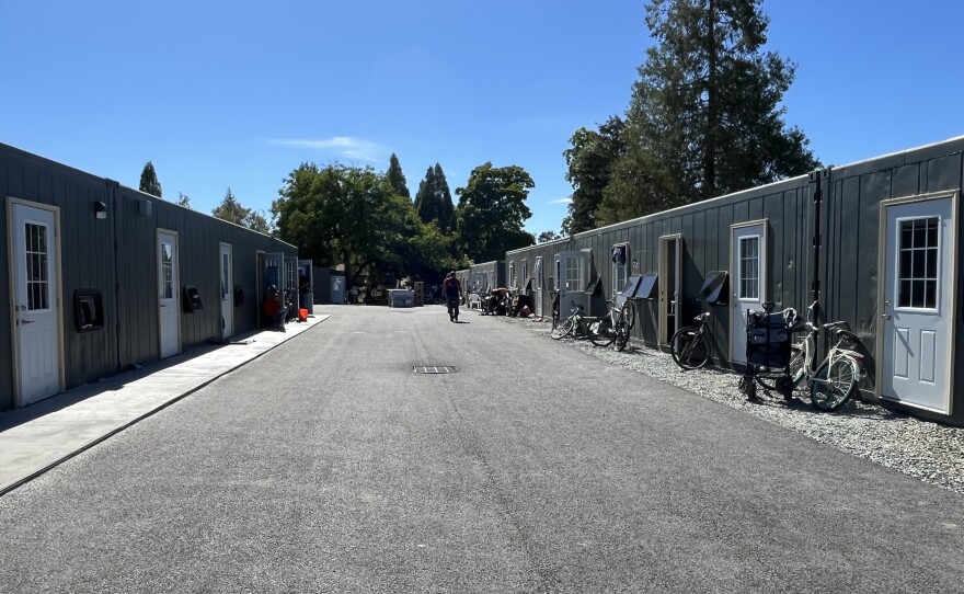 A wide driveway is lined with single-story shipping container homes. They are gray and have white doors and windows. 