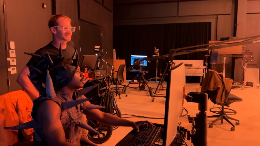 Student in spiky costume looking at a computer screen with teacher behind him.