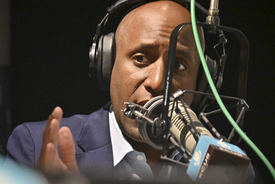 Close up image of a man gesturing with his left hand and talking at a microphone inside a radio studio.
