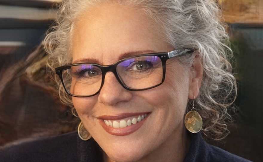 A smiling woman with curly grey hair is wearing eyeglasses,  round gold earrings and a blue zip-up fleece.