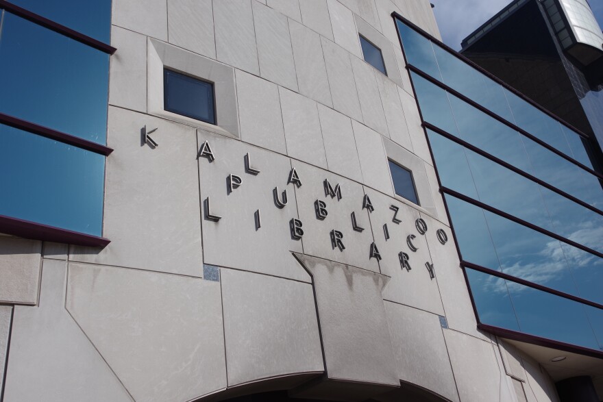 Silver letters on the side of a building read "Kalamazoo Public Library." The glass windows reflect the open blue sky on a bright sunny day.