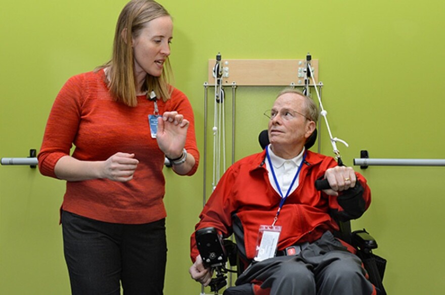 Head-on medium photo of a female physical therapist standing next to male patient in a wheelchair instructing him on the use of the pulley weights on the wall behind them.