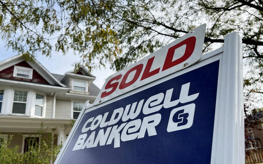 A blue and white Realtor sign that reads "Coldwell Banker" sits in front of a three-story home. Attached to the sign is a white sign with red letters that reads "Sold."