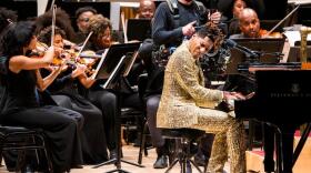 Composer & pianist Jon Batiste, in a suit of gold sequins, sits at the grand piano on the stage of Carnegie Hall in front of the Gateways Music Festival orchestra