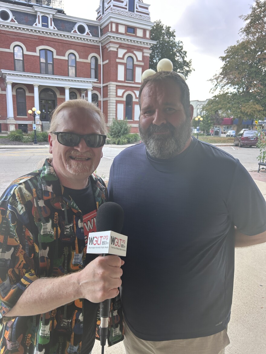 Two men stand outside in front of a historic brick building. One holds a WGUT 95.3 FM microphone and wears sunglasses and a shirt with guitars; the other wears a navy shirt and has a ping pong ball on his head.