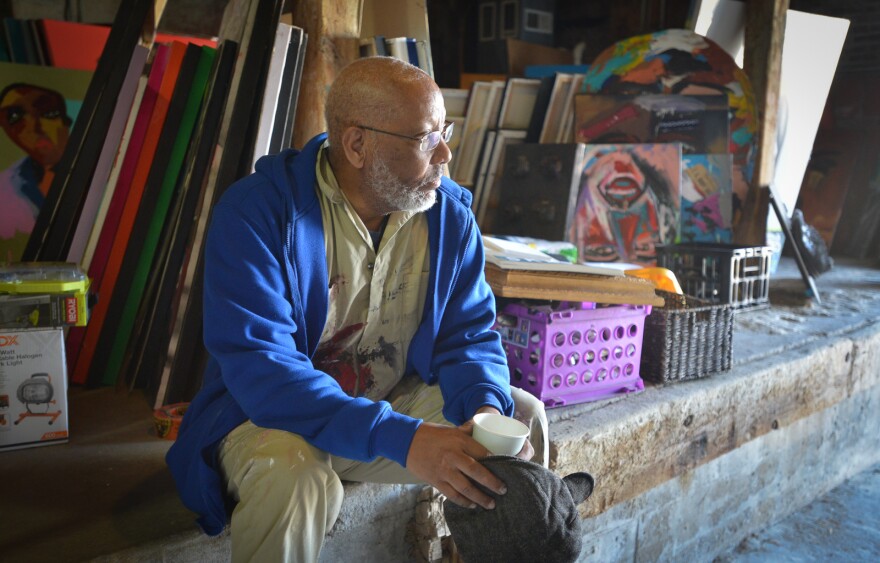 A man wearing tan coveralls and a blue jacket sits on the ledge of a concrete platform looking into the light that is beaming in from a garage door (not seen in photo). Behind him are many large painting canvases.