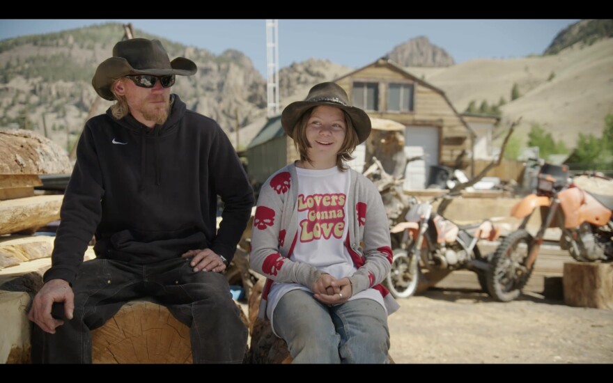 Dan and Lexy Mead sit on a wood block with a barn and several old motorcycles behind them. Dan has a reddish-blonde beard and sunglasses and wears a black hoodie and dark jeans. Lexy wears jeans, a wide-brimmed hat, and a shirt that says "lovers gonna love."